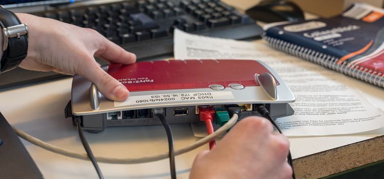 Das Bild zeigt die Hände einer Person, die eine Fritz-Box auf einem Schreibtisch manipuliert. Das Gerät hat ein rotes Oberteil und Kabel, die daran gesteckt sind. Im Hintergrund befindet sich eine Computertastatur und ein Notizbuch. Die Szene deutet darauf hin, dass die Person möglicherweise Verbindungen herstellt oder Fehler an dem Gerät behebt.