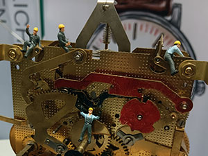 Ein Großuhrwerk, auf dem Miniaturfiguren mit gelbem Helm 'arbeiten'.