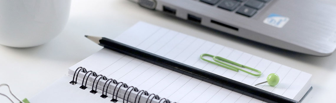 Ein Notizblock, auf dem ein Bleistift, eine grüne Büroklammer und ein Pin liegen. Im Hintergrund links oben leicht unscharf der Ausschnitt einer weißen Blumenvase. Rechts oben die Teilansicht eines Notebooks.
